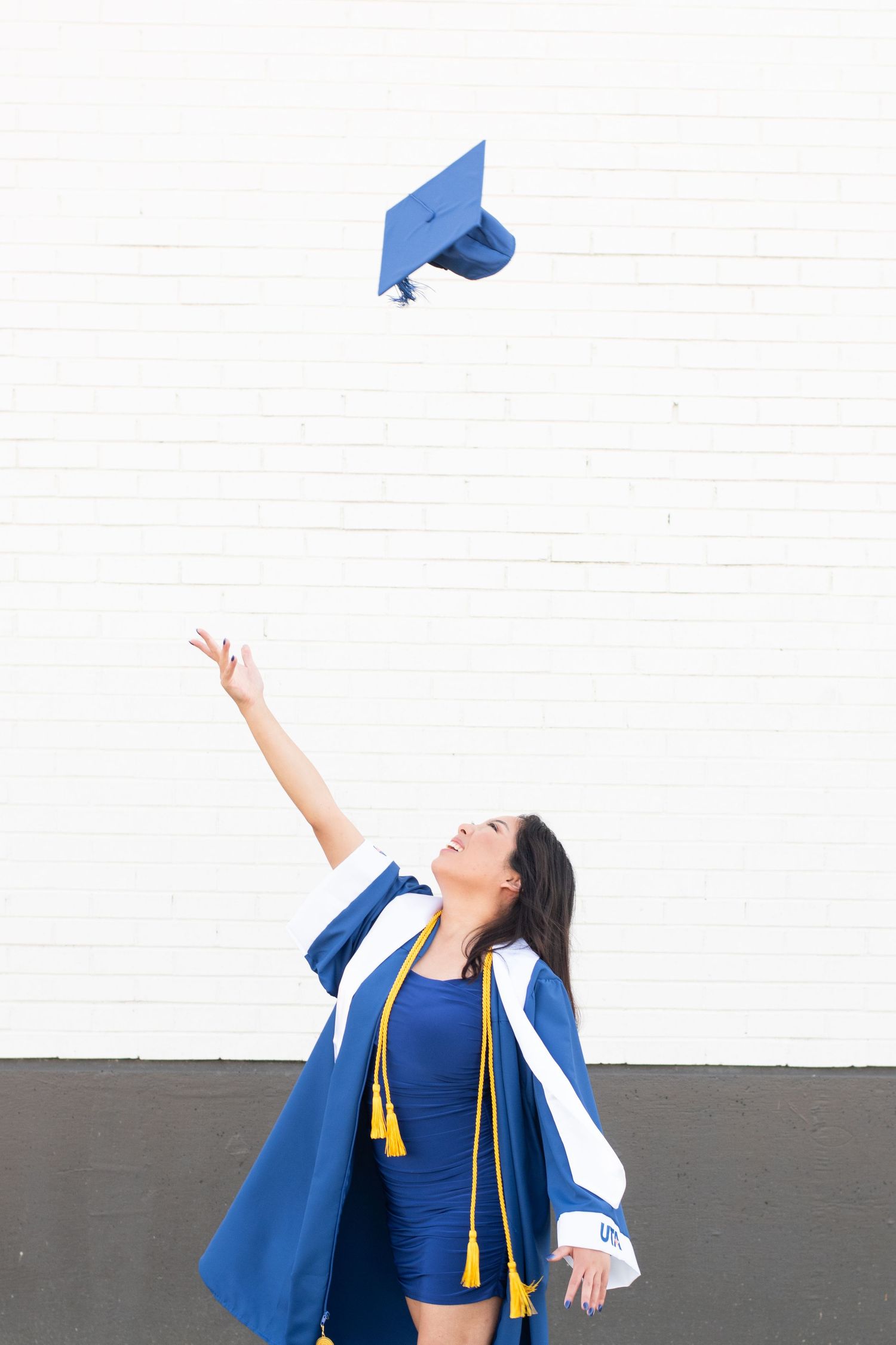 A graduate in a blue cap and gown throws their mortarboard cap into the air against a white wall background.