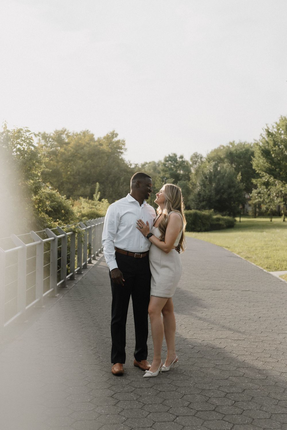 A romantic couple stands close together on a pathway with lush greenery and metal railings in the background.