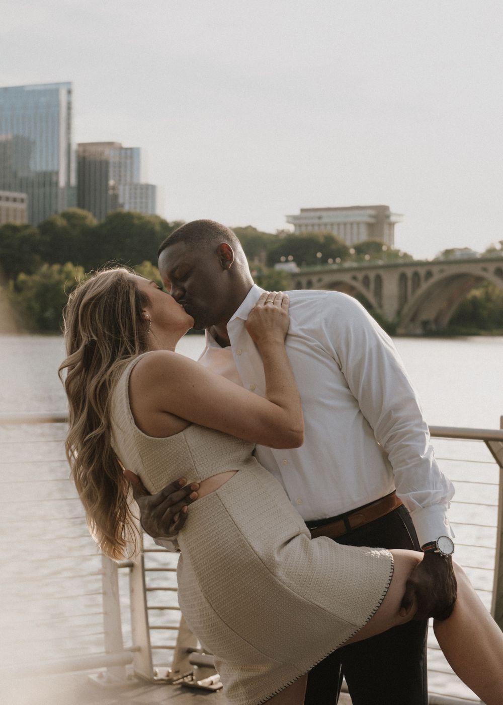 A romantic dip kiss captured on a waterfront walkway with city buildings in the background.