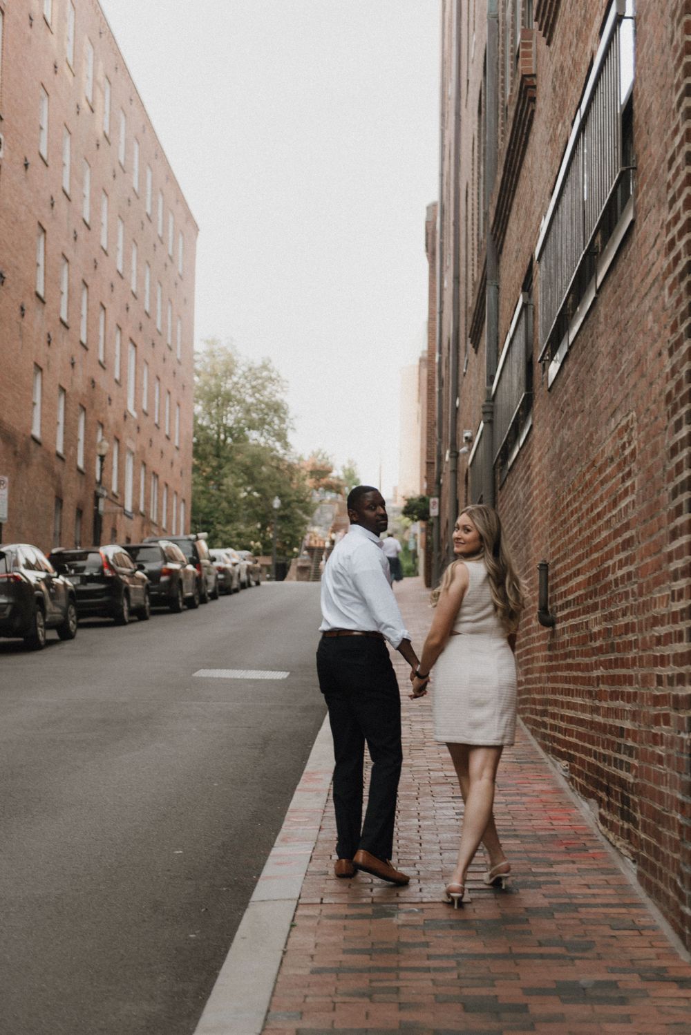 A couple walking hand in hand down a brick-lined city street with parked cars.