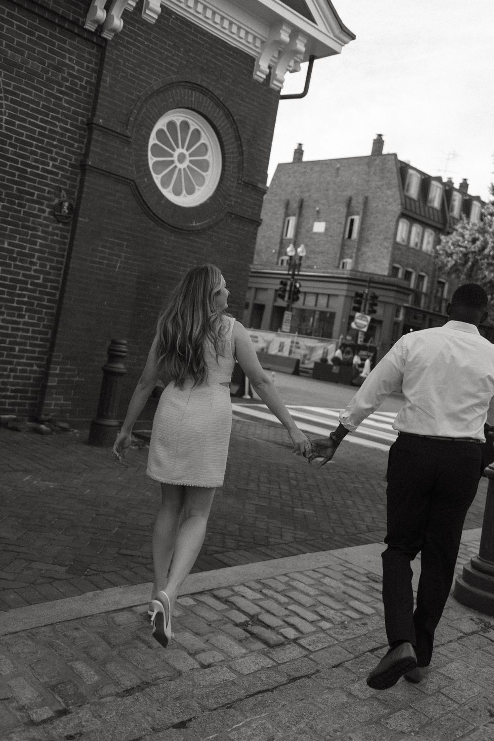 A couple walking hand in hand on cobblestone streets near a brick building with a round window.
