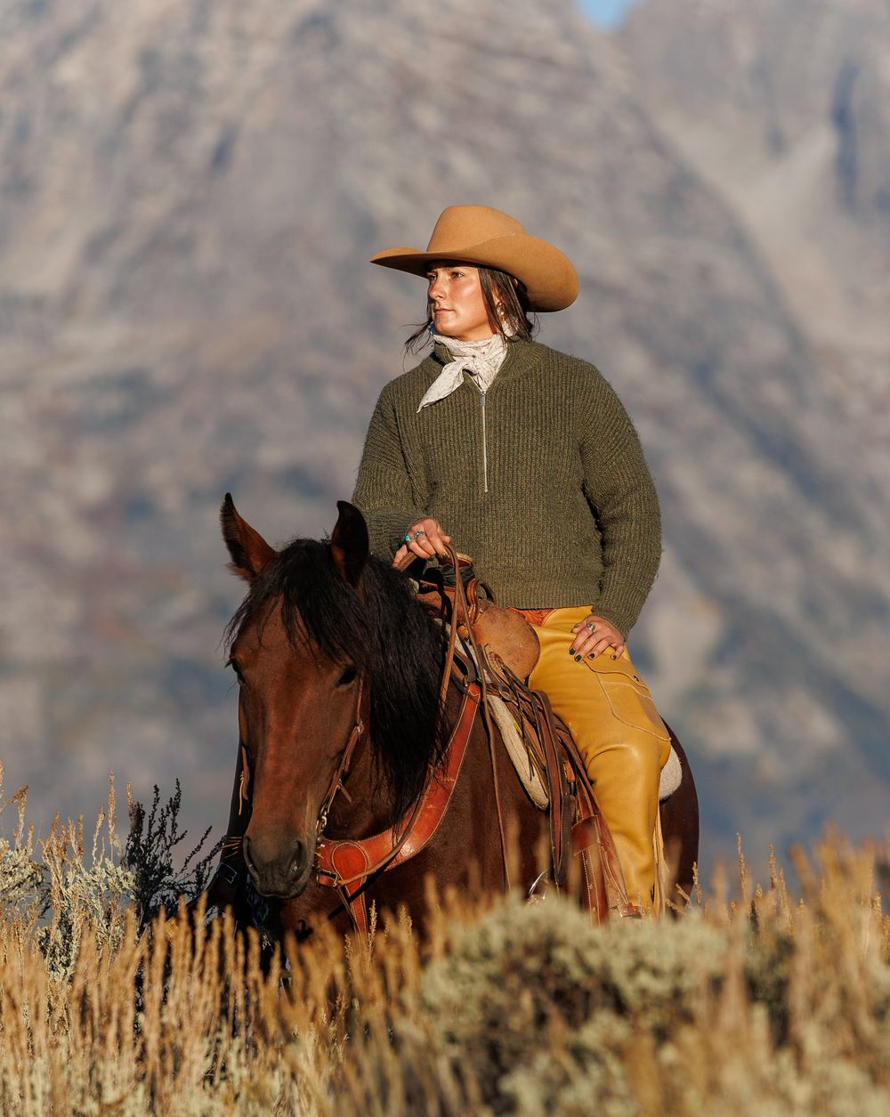 Riders on horseback traverse rugged mountain terrain with sagebrush in the foreground.