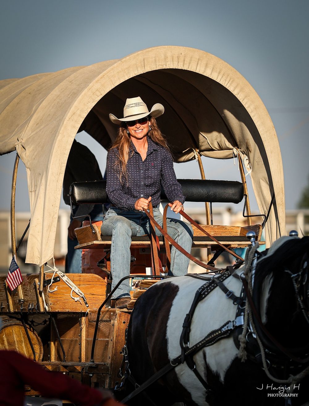 Cowgirl driving a covered wagon with horses in traditional western attire at sunset.
