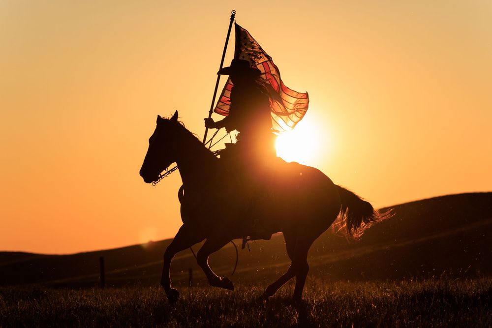 Silhouettes of horses and riders against dramatic orange sunset sky.