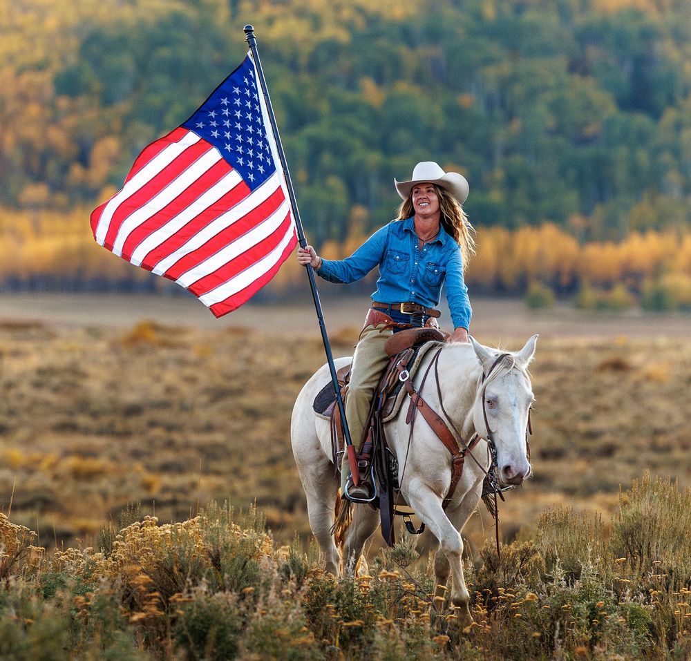 Riders carrying American flags on white horses gallop across an autumn field.