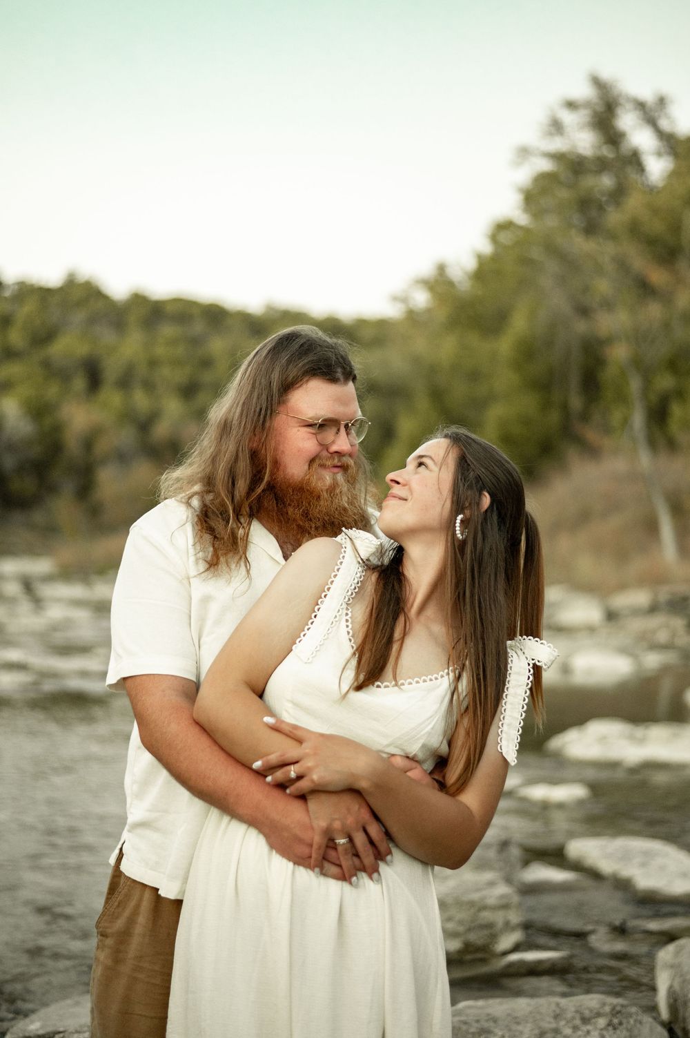 A couple in white outfits embraces outdoors, smiling at each other near a rocky riverbank with green trees behind.