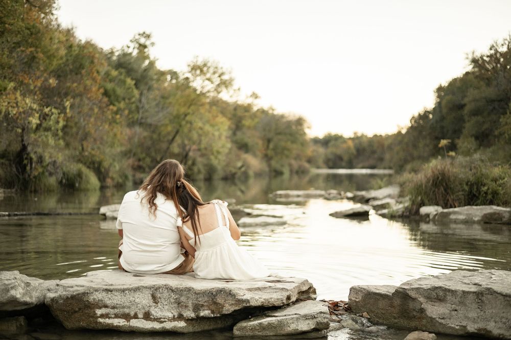 Couple in white sitting on rocks by a serene river surrounded by lush green trees in soft golden light.
