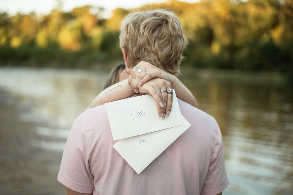 Couple in romantic embrace stands by tranquil lake at sunset with soft pink and golden lighting.