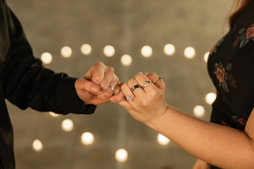 Couple holding pinkies with rings on fingers, candles glowing in heart shape in background, romantic proposal moment.