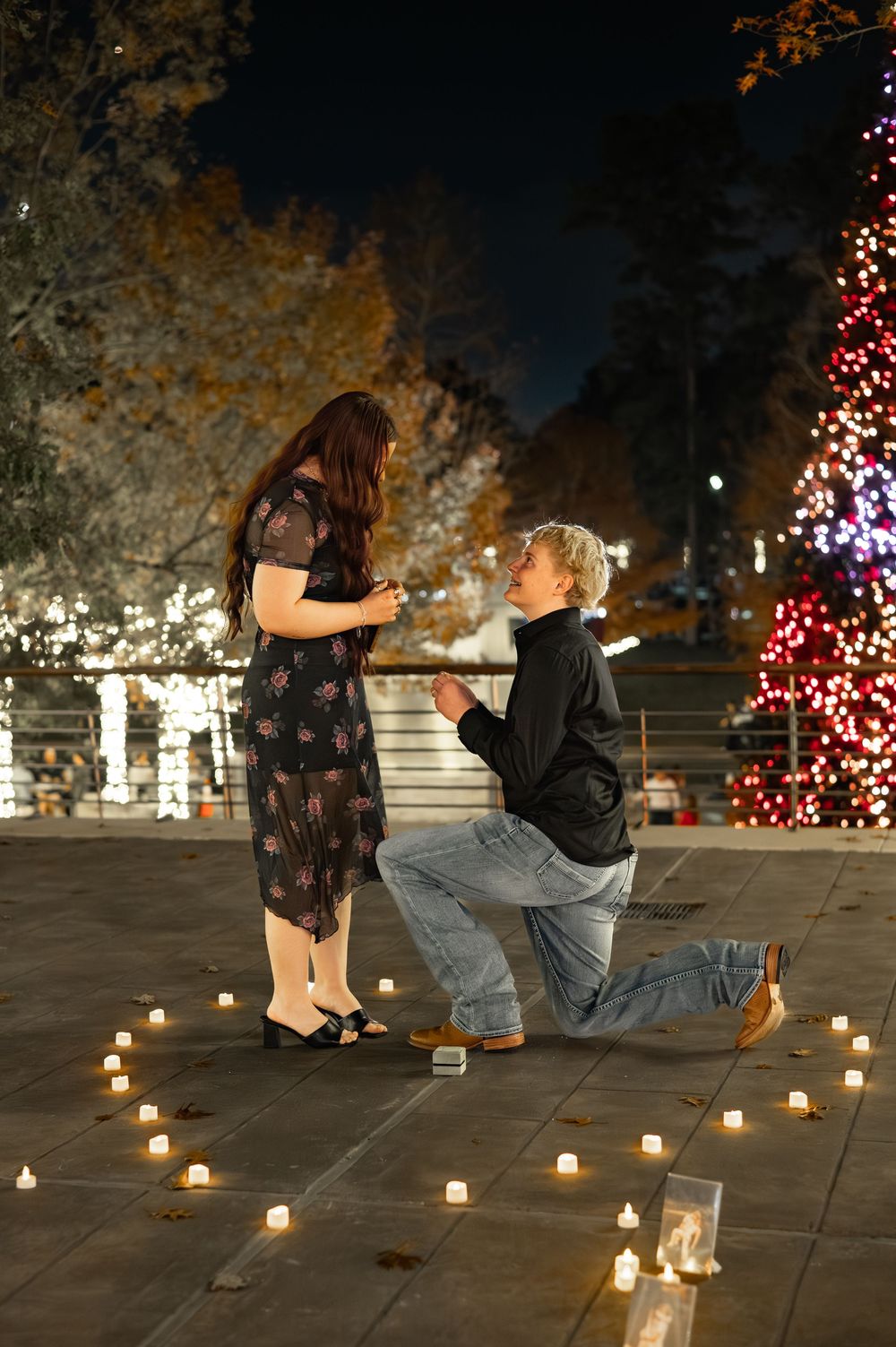 Person proposing on one knee with candles in heart shape, Christmas tree lights glowing at night outdoor setting.