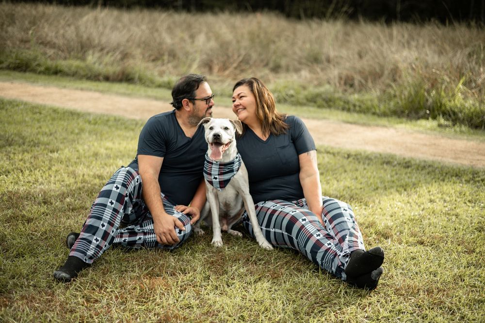 Couple in matching plaid pajamas sitting on grass with white dog between them, smiling at each other outdoors.