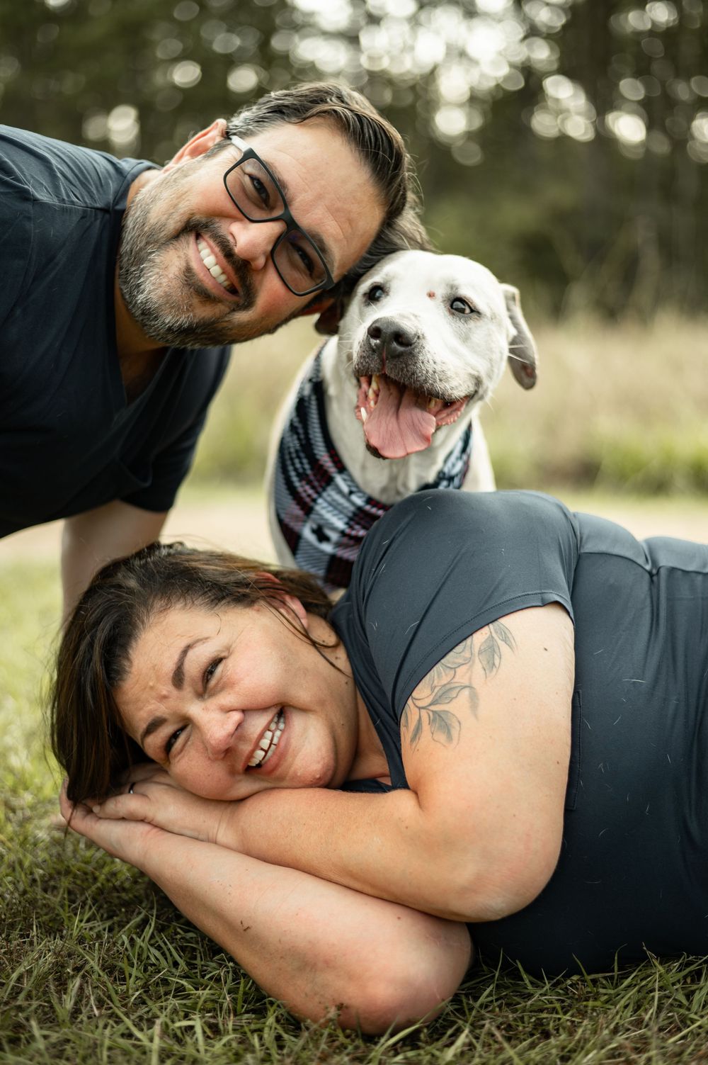 A smiling couple poses outdoors on grass with their happy white dog wearing a bandana.