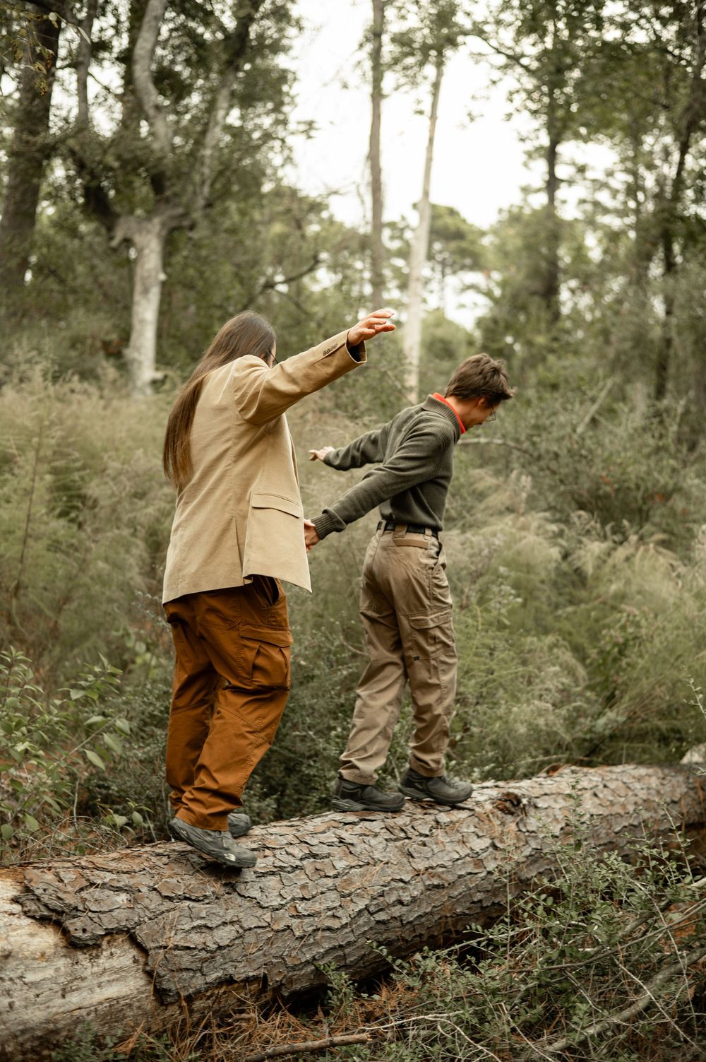 Two people balancing on a fallen log in a lush forest, wearing earth-toned outdoor clothing.