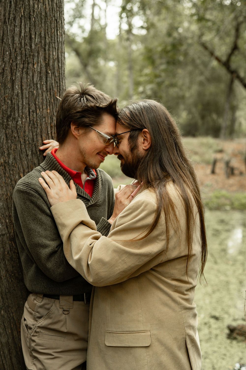 Romantic couple embracing by a tree in a forest, wearing glasses and casual jackets, sharing an intimate moment.