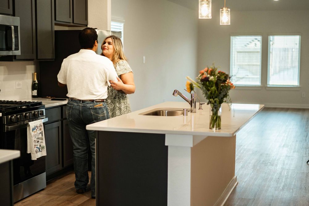 Couple embracing in a modern kitchen with white countertops, dark cabinets, and fresh flowers on the island.