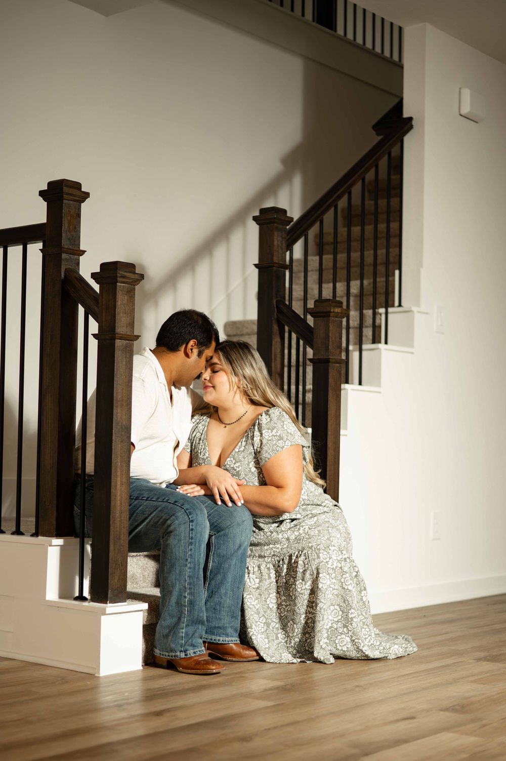Couple sitting on staircase steps, leaning foreheads together romantically, in a bright modern home with hardwood floors.