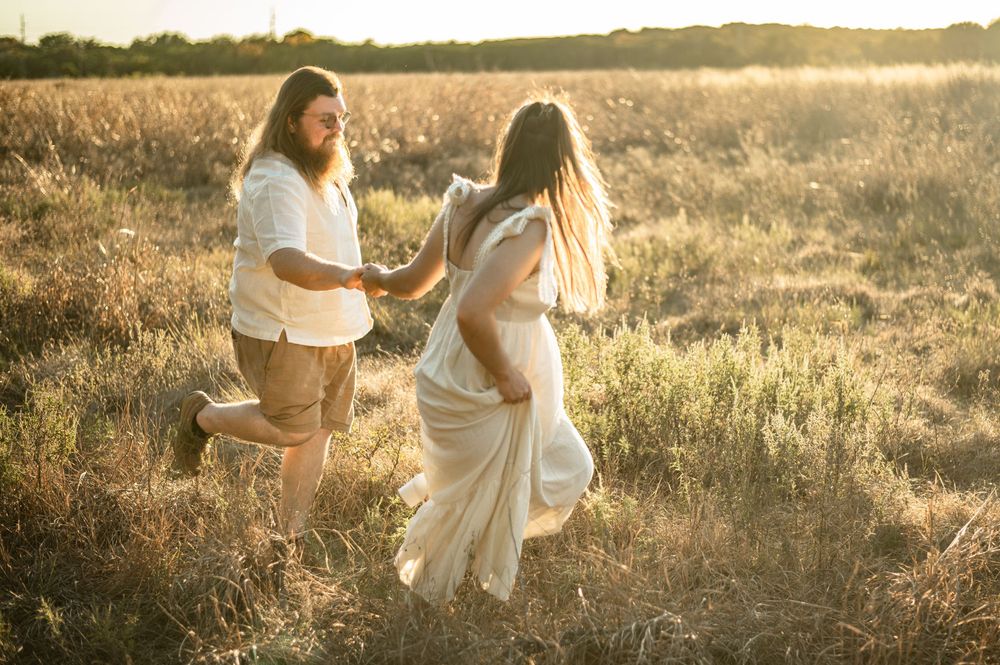 Couple dancing joyfully in golden sunlit field, woman in flowing cream dress, man in white shirt and shorts.