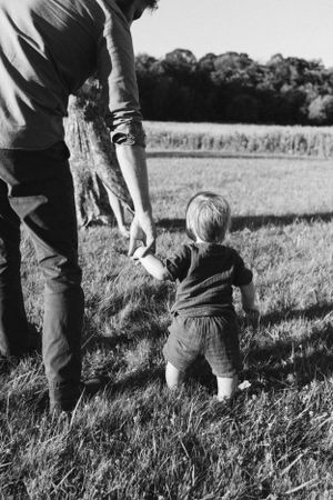 Parent and child hold hands while walking through grassy field in black and white.