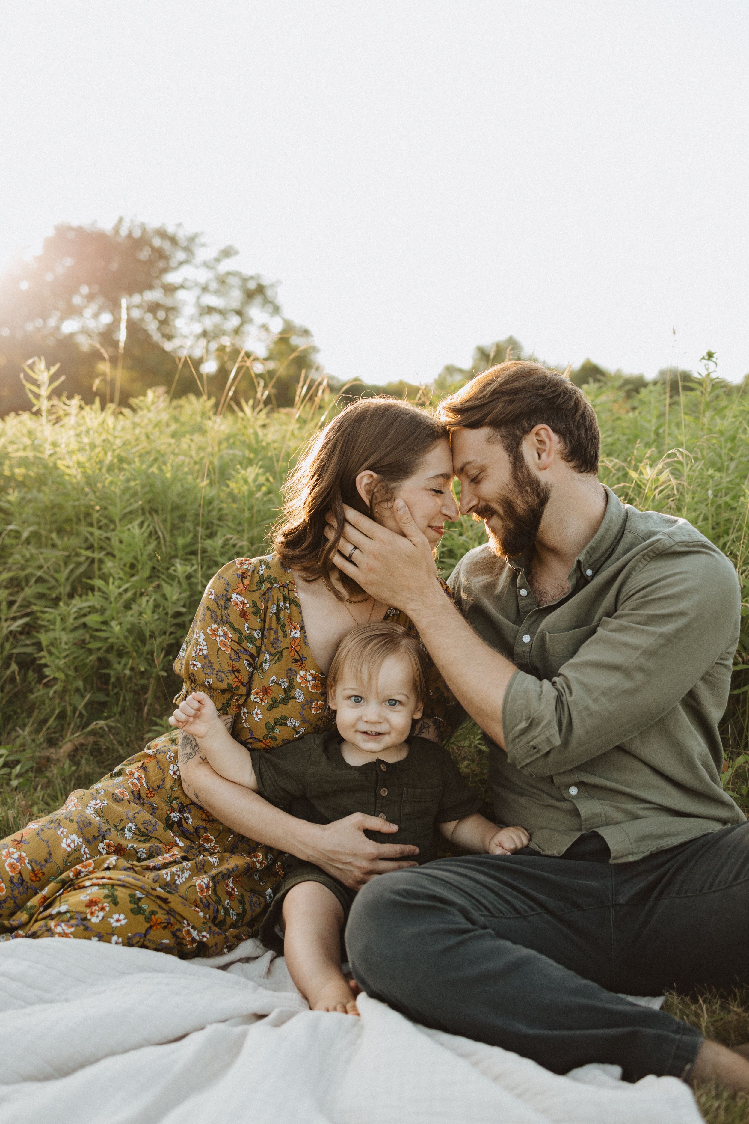 Family sits on blanket in golden field at sunset sharing intimate moment together.