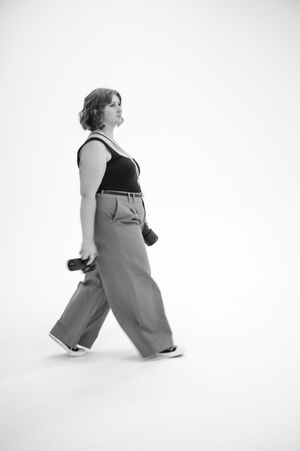 A person wearing wide-leg pants and a tank top walks sideways in a minimalist black and white studio setting.