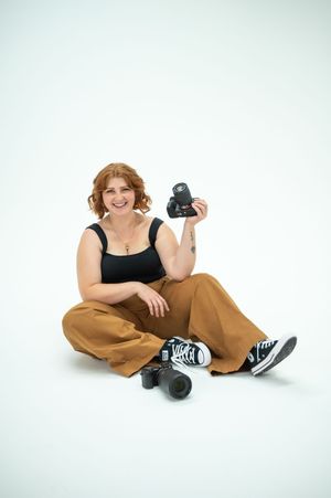 A photographer sits casually on white floor with camera gear while wearing tan trousers and black tank top.