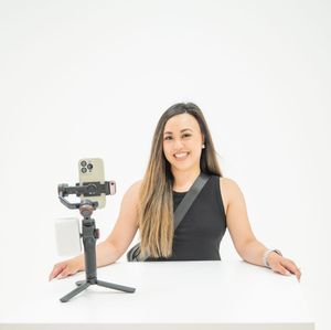 Content creator filming on camera mounted to mini tripod on white desk in studio against light background.