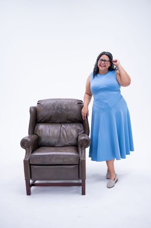 A brown leather loveseat and someone wearing a light blue dress pose in a studio setting against a white background.