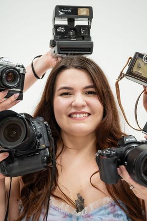 A lifestyle image showing a photographer surrounded by various cameras including vintage and modern DSLR equipment.