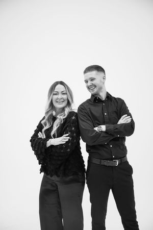 A couple wearing black outfits poses together in a fun and playful way against a white background.