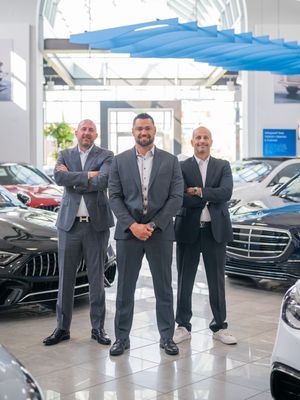 Professional car dealership staff in suits standing together in a modern showroom with luxury vehicles on display.