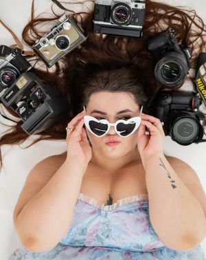 A creative flat lay portrait with a person lying among vintage cameras wearing heart-shaped white sunglasses.