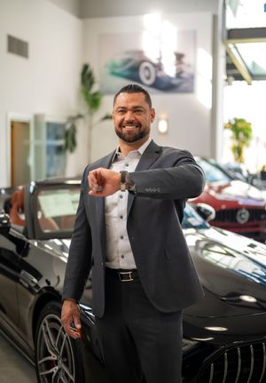 A well-dressed person in a black suit poses confidently next to luxury vehicles in an upscale showroom setting.