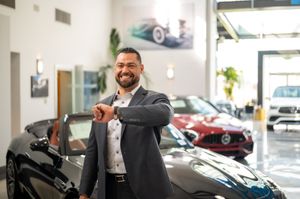 A person in a suit smiles while gesturing in a luxury car showroom with vehicles displayed in the background.
