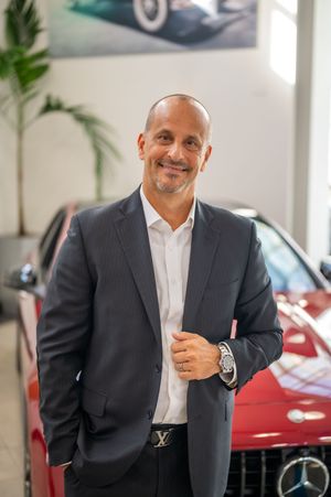 Business professional in dark suit stands with positive expression in front of luxury vehicles at car dealership.