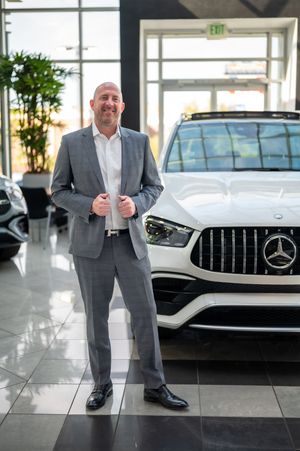 Professional in a gray suit poses confidently in a modern Mercedes-Benz showroom with luxury vehicles on display.
