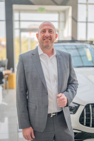 Professional in a gray suit and white shirt smiles warmly in a bright car dealership showroom.
