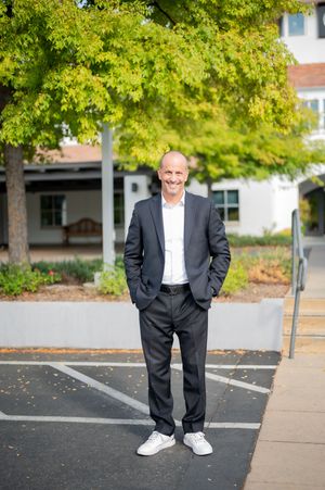 A person in a dark suit and white sneakers stands in an outdoor parking lot with green trees in the background.