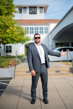 A person in a dark suit and sunglasses stands outside a modern building with Mercedes-Benz signage.