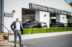 Professional walking outside a modern Mercedes-Benz dealership building with white exterior walls and landscaping.