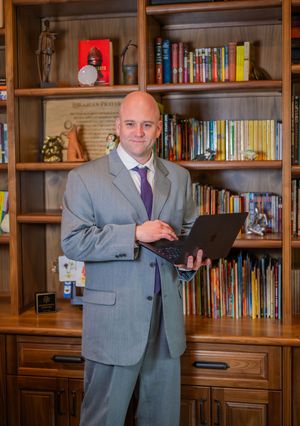 Professional in a gray suit stands in front of wooden bookshelves in an office setting.