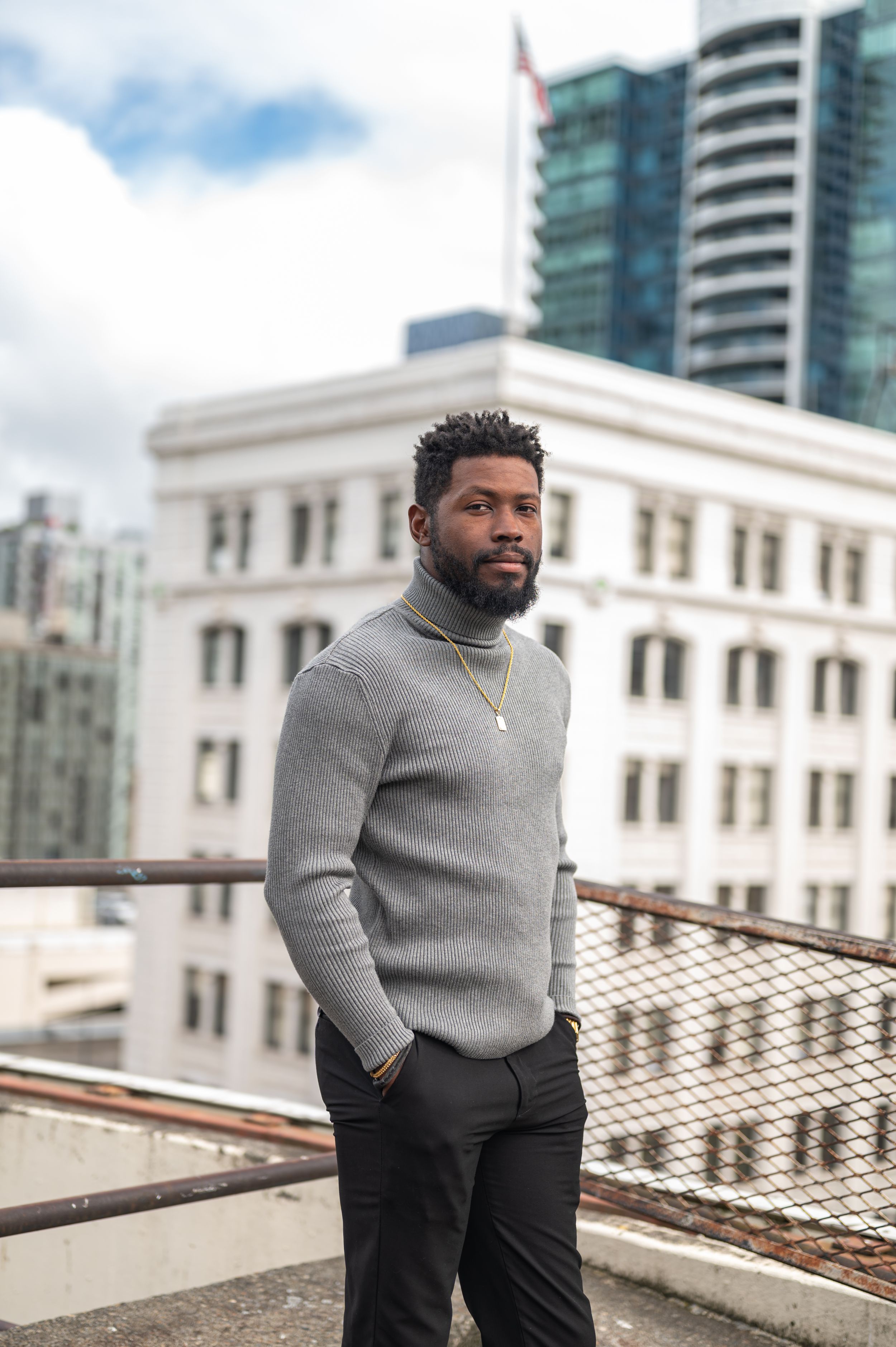 Urban fashion portrait featuring grey sweater and dark pants with city buildings in background.