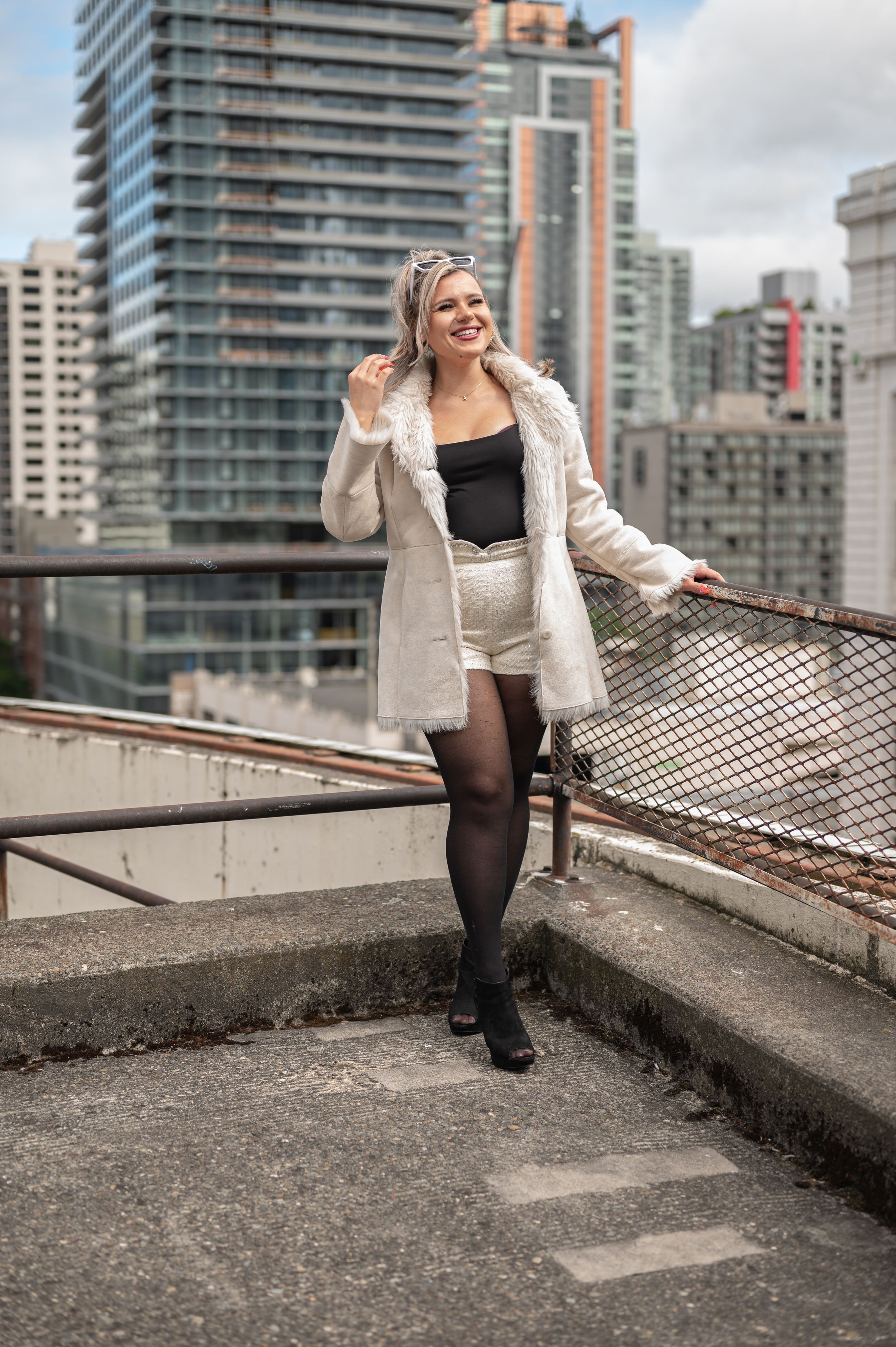 Fashion model wearing cream coat and shorts poses on urban rooftop with city skyline backdrop.