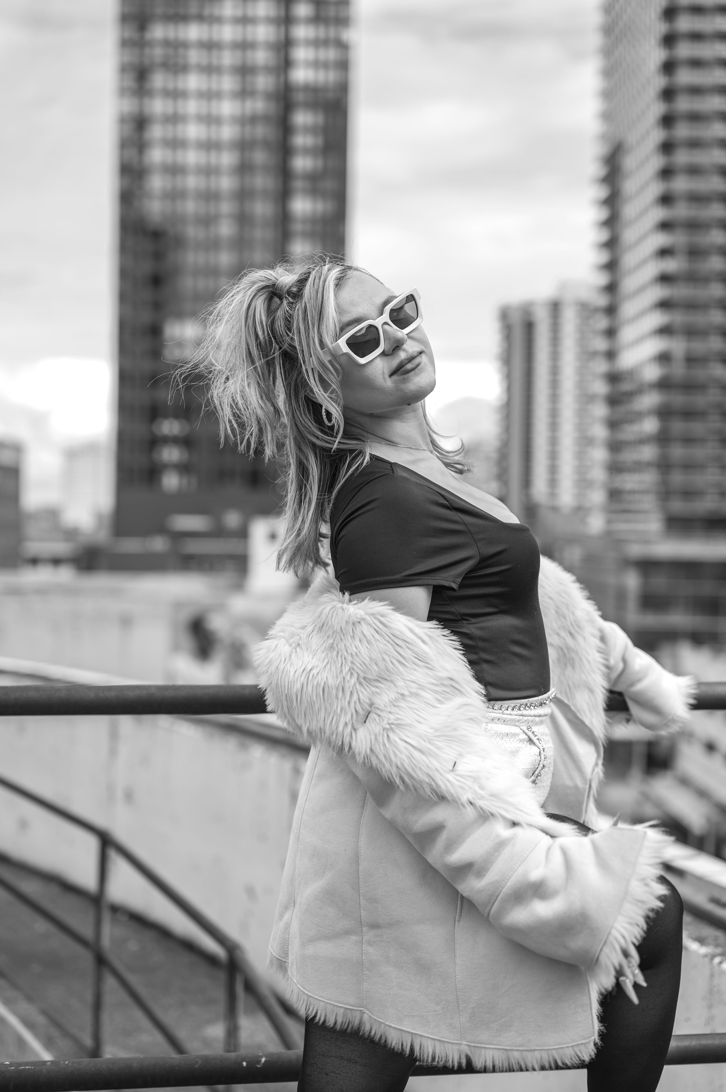 Woman in fur jacket poses against urban skyline in black and white photography.