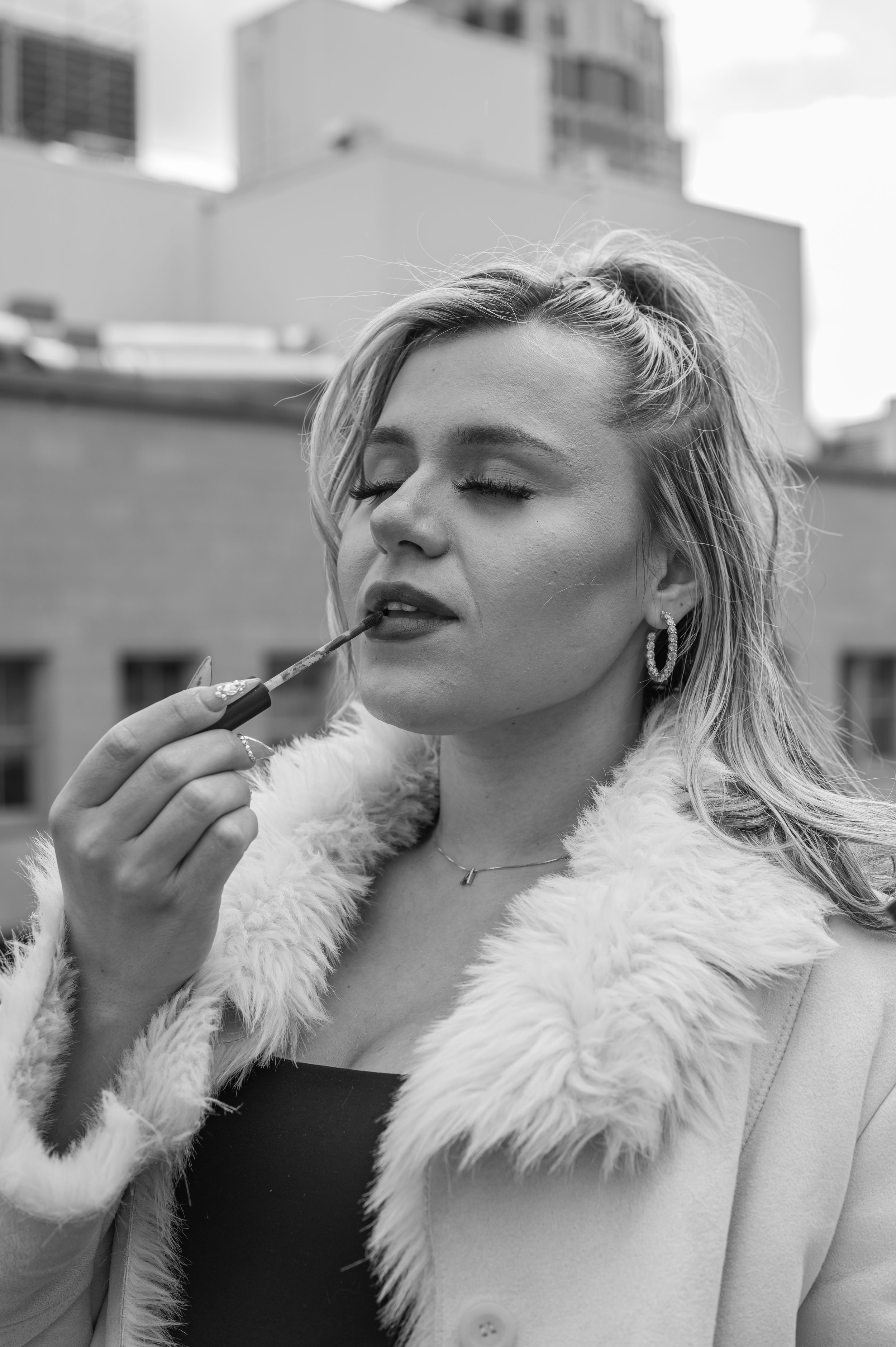 Black and white portrait of woman in fur coat applying lipstick outdoors.