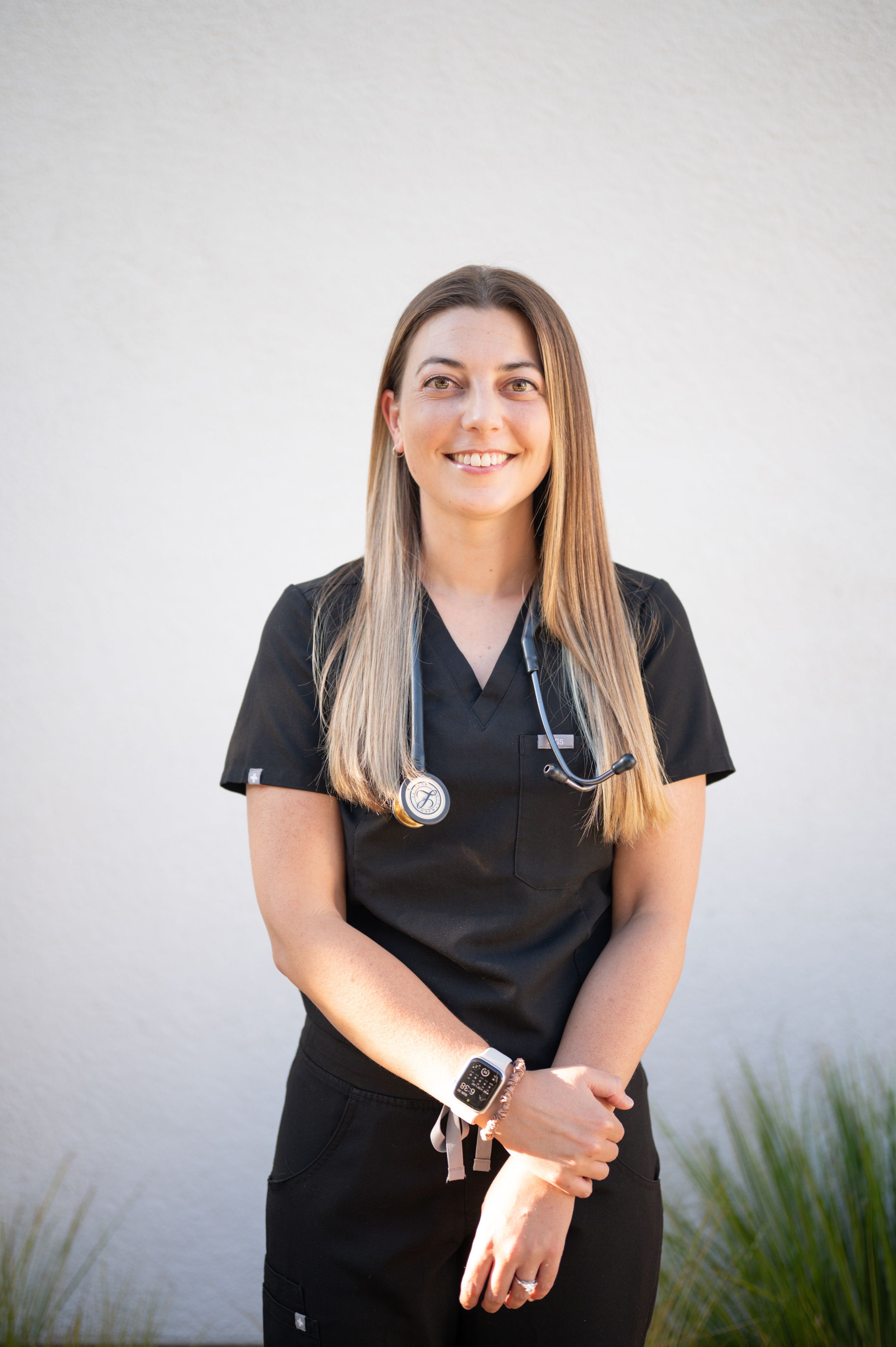 Healthcare worker in black scrubs with stethoscope poses against white wall.