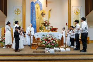 Religious ceremony with altar servers and clergy gathered around decorated altar with blue and white floral arrangements.