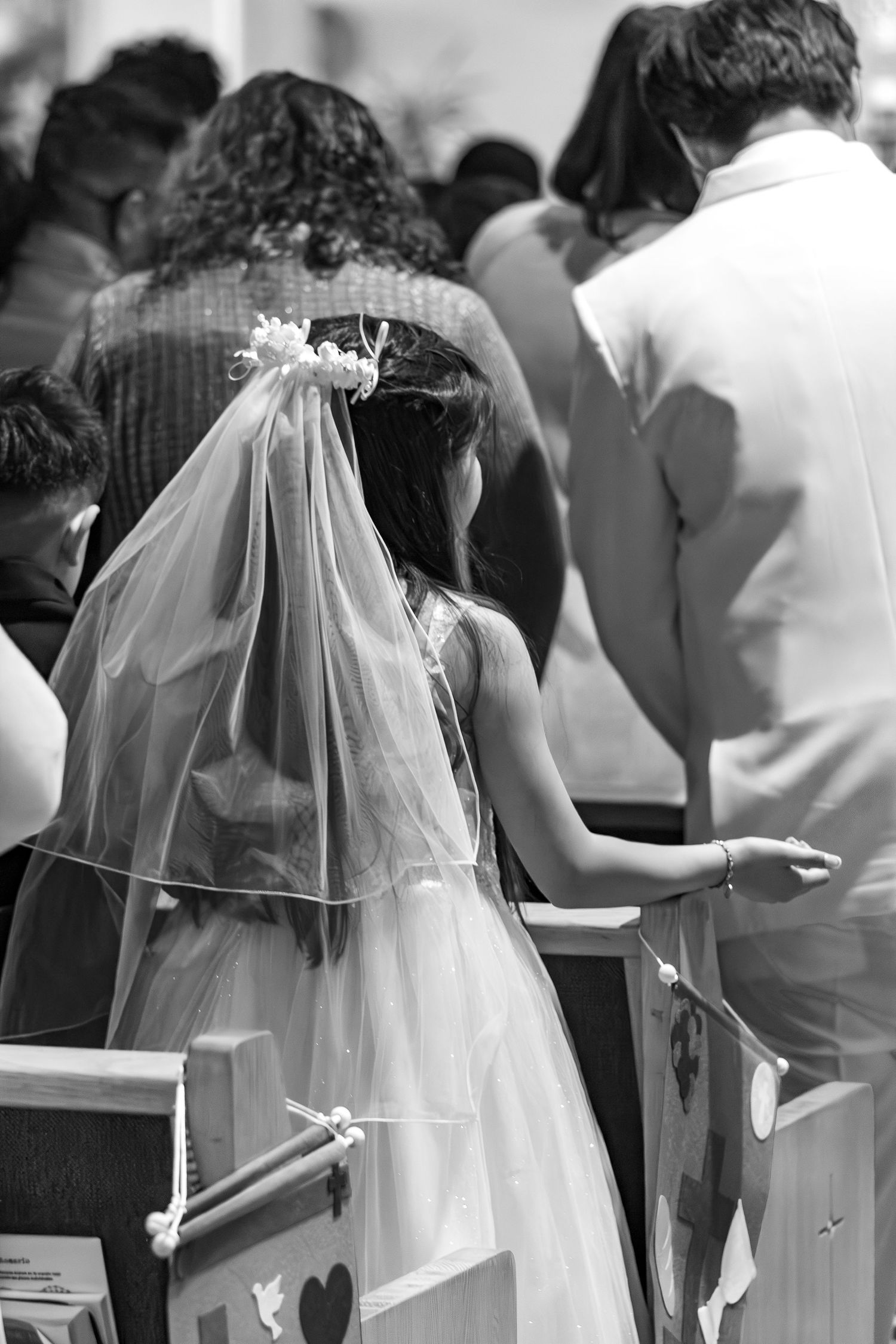 A bride in a white dress and veil stands among a crowd at a wedding ceremony, shown in black and white photography.