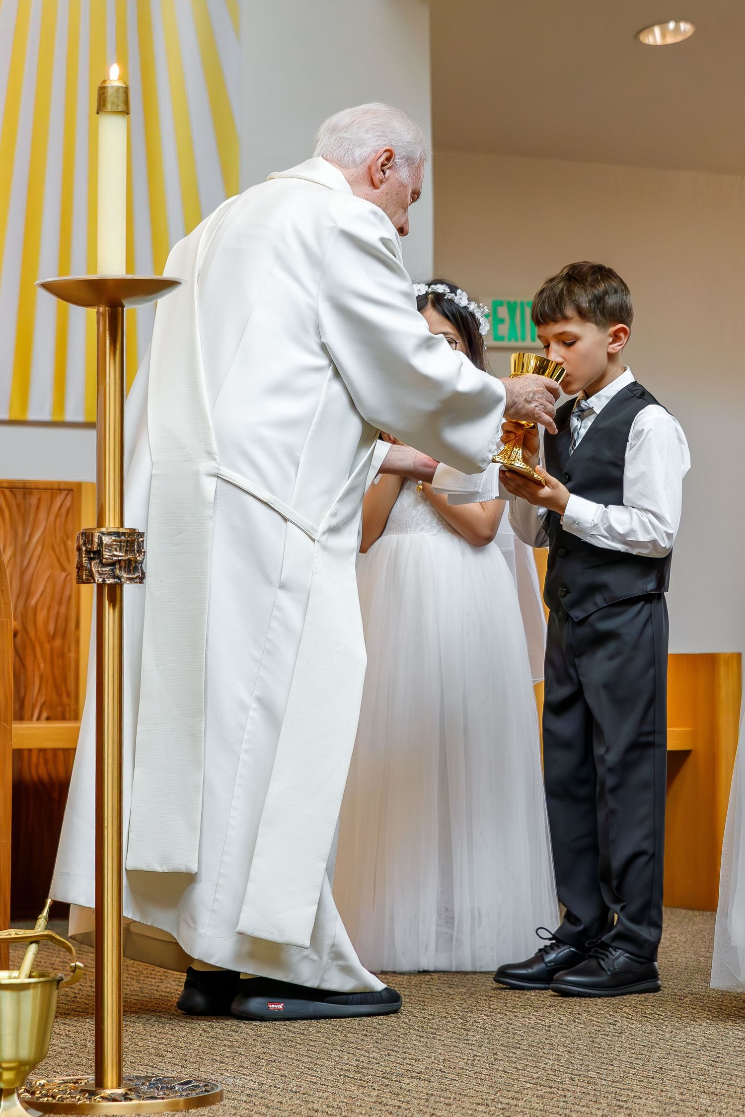 A religious ceremony takes place inside a church as two people interact at the altar with yellow decorative stripes on the wall.