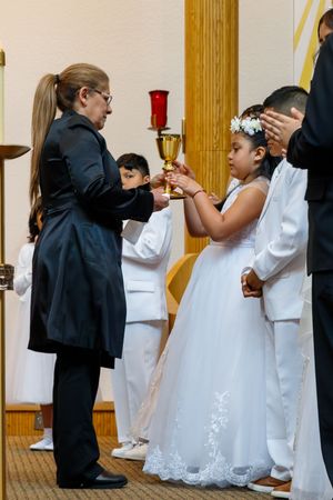 Children in white dresses and dark suits stand in line at church receiving communion during a religious ceremony.