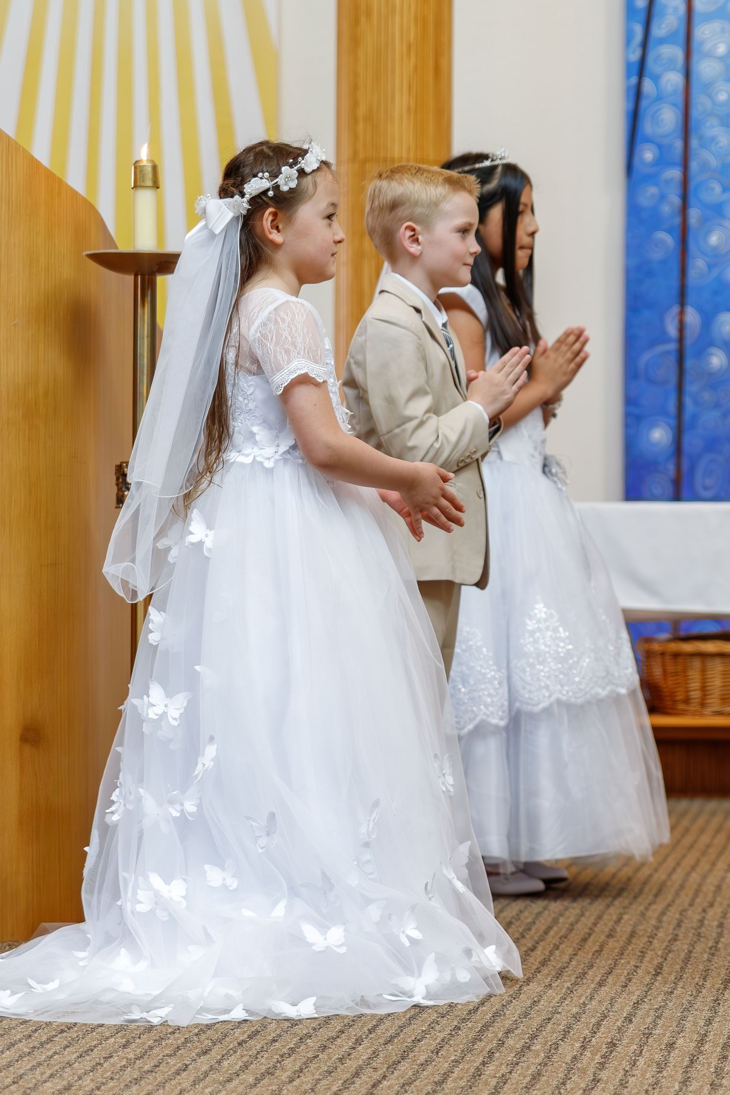 Children in formal attire stand at the altar during a religious ceremony with white dresses and formal suits.